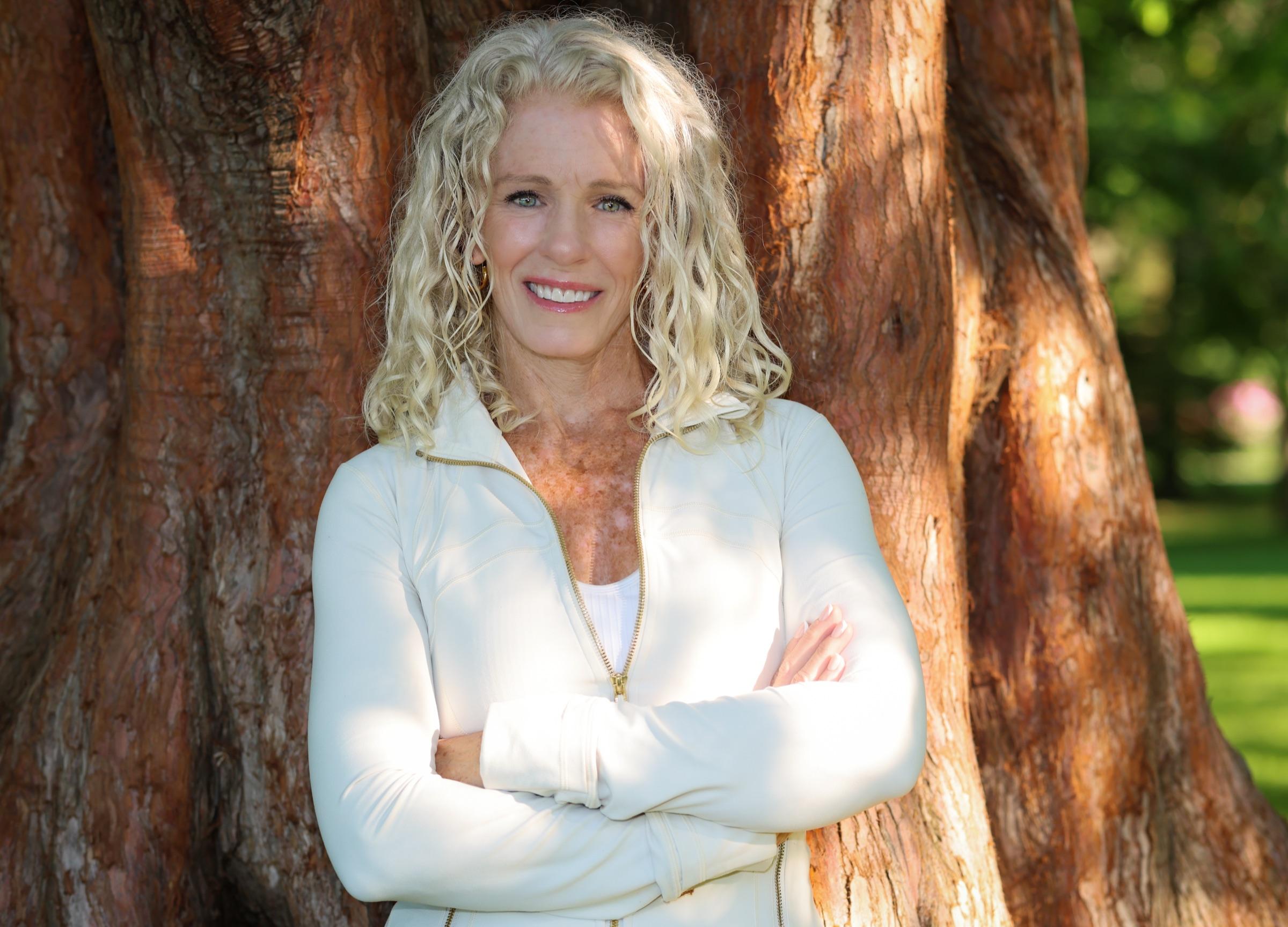 Stephanie Colwell smiling warmly, arms gently folded, standing in front of a large cedar tree