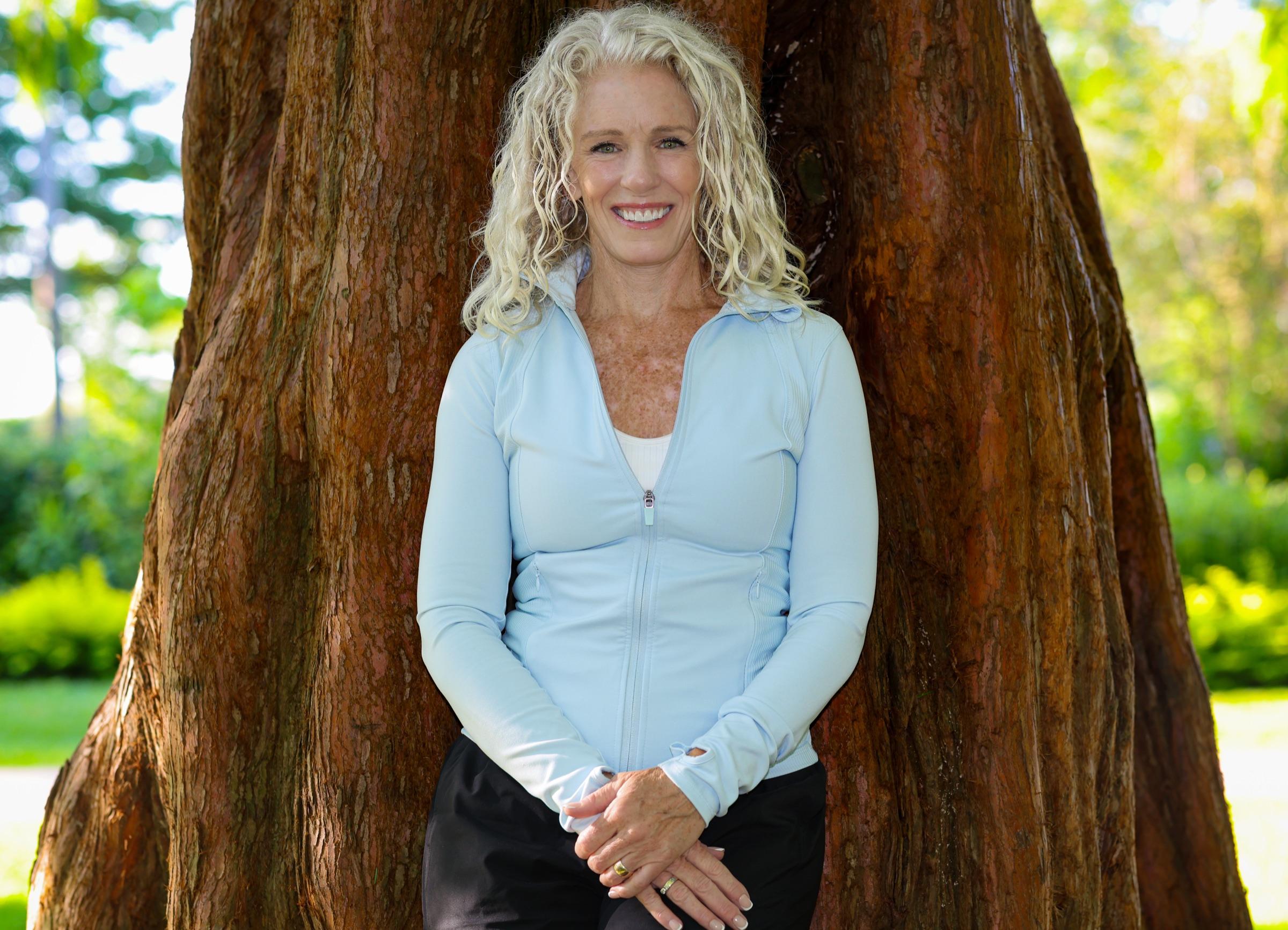 Stephanie Colwell in a pale blue jacket standing at the base of a broad cedar tree